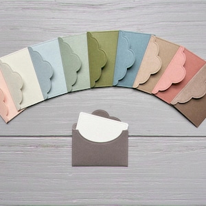 May include: A collection of colorful, scalloped-edge card holders in various pastel shades, arranged in an arc. A single holder with a white card is in the foreground. The holders are on a gray wooden surface.