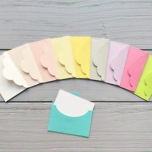 May include: A selection of pastel-coloured mini envelopes with scalloped edges, arranged in an arc. A teal envelope holds a white card. The envelopes are on a grey wooden surface, suitable for stationery or gift packaging.