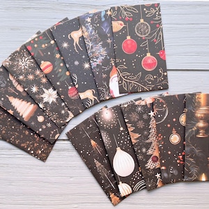 May include: A collection of rectangular paper envelopes featuring various dark-themed holiday designs, including illustrations of Christmas trees, ornaments, reindeer, snowflakes, and lit candles, arranged in a fan shape on a light grey surface.