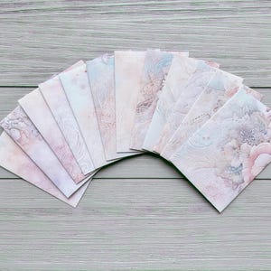 May include: A fan-shaped arrangement of ten rectangular envelopes featuring a soft, muted colour palette of pale pink, dusty blue, and cream. Each envelope displays a delicate, intricate floral and swirling filigree pattern printed on the paper.