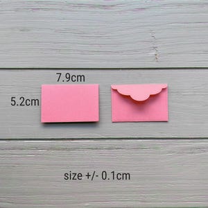 May include: Two small pink paper envelopes, one closed and one open with a scalloped flap. The dimensions are labeled as 7.9 cm by 5.2 cm, with a size tolerance of +/- 0.1 cm. The envelopes are rectangular and feature a smooth, matte finish.