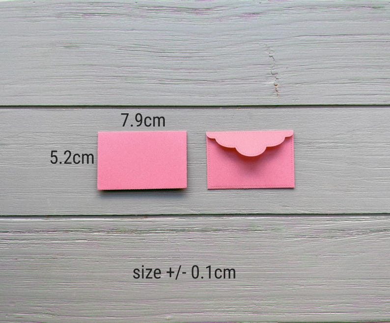 May include: Two small pink paper envelopes, one closed and one open with a scalloped flap. The dimensions are labeled as 7.9 cm by 5.2 cm, with a size tolerance of +/- 0.1 cm. The envelopes are rectangular and feature a smooth, matte finish.