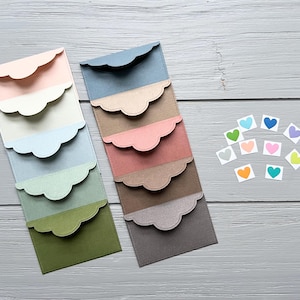 May include: Two vertical stacks of rectangular paper envelopes with scalloped flaps in a gradient of colors including pink, cream, blue, green, brown, and gray, accompanied by ten small square stickers featuring colorful heart illustrations.