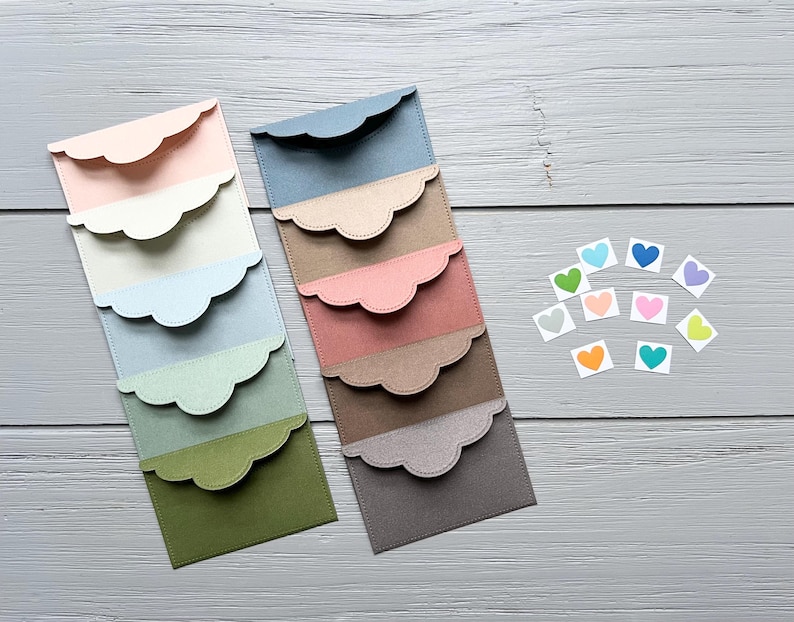 May include: Two vertical stacks of rectangular paper envelopes with scalloped flaps in a gradient of colors including pink, cream, blue, green, brown, and gray, accompanied by ten small square stickers featuring colorful heart illustrations.