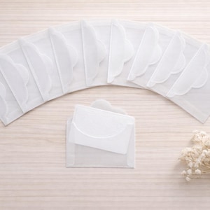 May include: A collection of white, semi-transparent pouches with a scalloped edge, arranged in an arc. A smaller pouch is in the centre. Dried white flowers are in the bottom right. The pouches are likely made of a lightweight material.