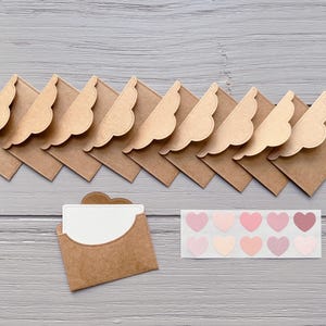 Puede incluir: Un conjunto de sobres de papel kraft con solapas festoneadas, dispuestos sobre una superficie de madera gris. Un sobre está abierto, revelando una tarjeta blanca. También hay una tira de pegatinas en forma de corazón en varios tonos de rosa.