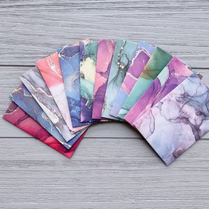 May include: A collection of rectangular envelopes arranged in a fan shape, each featuring a unique abstract alcohol ink design with swirling patterns in shades of pink, purple, blue, green, and gold, creating a vibrant and colourful display.