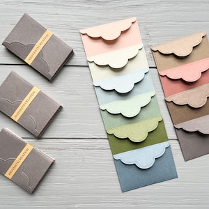 May include: Three grey rectangular card holders with yellow bands labeled ANDYBATS CREATION, alongside two vertical stacks of rectangular envelopes featuring scalloped flaps in various pastel and earth-toned colors including pink, cream, blue, and green.