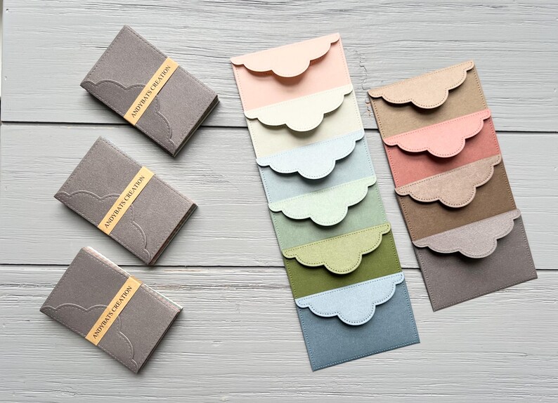 May include: Three grey rectangular card holders with yellow bands labeled ANDYBATS CREATION, alongside two vertical stacks of rectangular envelopes featuring scalloped flaps in various pastel and earth-toned colors including pink, cream, blue, and green.