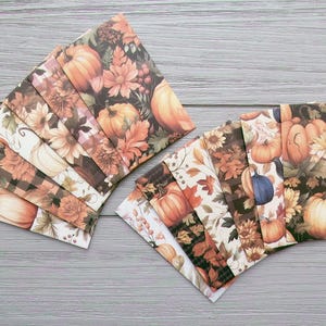 May include: Two stacks of decorative paper envelopes with autumnal patterns. The designs feature pumpkins, sunflowers, and autumn leaves in shades of orange, brown, and cream. The envelopes are arranged on a light grey wooden surface.