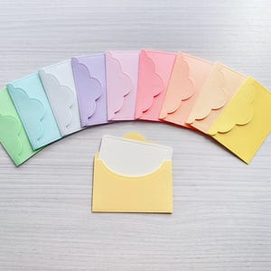 Op de afbeelding: Een verzameling van tien kleine enveloppen in een spectrum van pastelkleuren waaronder groen, blauw, wit, lavendel, roze, perzik en geel, elk met een geschulpte klep, met één gele envelop op de voorgrond die een witte kaart bevat.