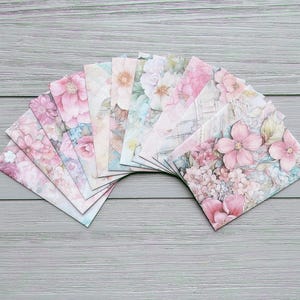 May include: A selection of floral-patterned stationery envelopes fanned out on a grey wooden surface. The envelopes feature various watercolour designs of pink and peach flowers, with hints of blue and green. The stationery is ideal for sending letters or notes.