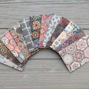 Op de afbeelding: Een verzameling decoratieve papieren enveloppen met kleurrijke, patroonontwerpen. De enveloppen zijn in een waaier gerangschikt en tonen een verscheidenheid aan geometrische en bloemmotieven in tinten rood, blauw, bruin en beige. Ze liggen op een grijze houten ondergrond.