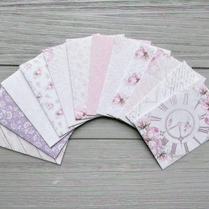 May include: A collection of rectangular paper envelopes arranged in a fan shape. The envelopes feature various designs including light purple floral patterns, soft pink textures, script writing, and a prominent clock face surrounded by pink roses.