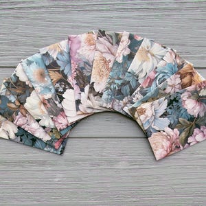 May include: A collection of rectangular fabric swatches arranged in a fan shape, each featuring a detailed floral pattern with large blossoms in shades of dusty pink, muted blue, cream, and dark green against a dark, moody background.