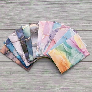 Puede incluir: Una colección de hojas de papel de arte abstracto y colorido abanicadas sobre una superficie de madera. Las hojas muestran una variedad de diseños estilo acuarela en tonos de azul, rosa, morado y dorado. El papel está dispuesto en semicírculo.