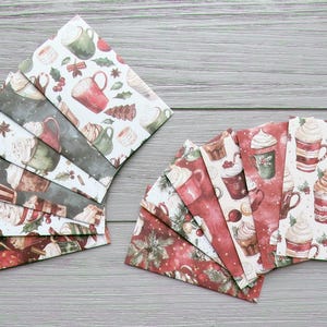 May include: A collection of rectangular paper envelopes featuring a repeating watercolour pattern of mugs filled with whipped cream, cinnamon sticks, and holly leaves. The envelopes are arranged in two fan-shaped clusters, displaying white, dark green, and red backgrounds.