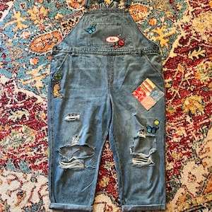 Puede incluir: Pantalón vaquero azul claro con detalles desgastados y varios parches bordados de colores, incluyendo mariposas, una flor, un tigre y una cara sonriente. El pantalón tiene tirantes ajustables y un parche con el nombre "Kyle" en la parte delantera.