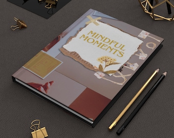 May include: A closed journal with a black cover and a collage of images on the front. The journal has the words "Mindful Moments" printed in gold on the front. The journal is sitting on a brown surface with two pencils and a gold metal decorative stand.
