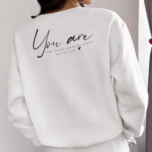 以下が含まれることがあります： 背面に「You are loved. Strong. Important. Worthy. Beautiful」というテキストと小さなハートが書かれた白いスウェットシャツです。