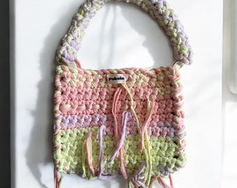 May include: A pastel pink, yellow, and purple crocheted tote bag with a crocheted handle and fringe details. The bag has a small label that says "Pokolo".