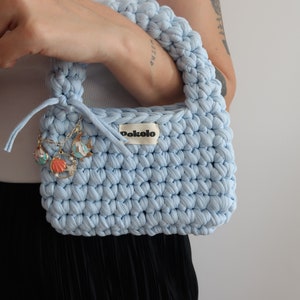 May include: A light blue crocheted handbag with a small white label that says "Dokolo" and gold charms attached to the handle.