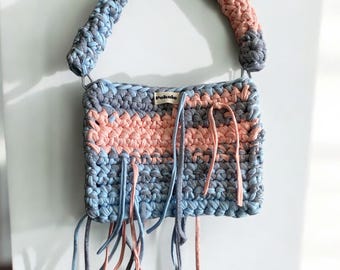 May include: A small, crocheted handbag with a pink, blue, and gray striped design. The bag has a single strap and a label that reads "Pokolo".