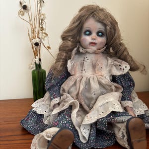 May include: A doll with long wavy brown hair and glowing blue eyes, wearing a white eyelet lace pinafore over a dark blue floral print dress with ruffled sleeves and lace trim, featuring a pale face with dark shading around the eyes and lips.