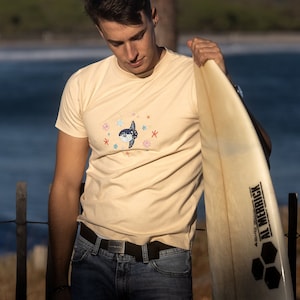 May include: A person wearing a light beige t-shirt with a blue and white fish graphic and a black belt. They are holding a white surfboard with black text that says "Al Merrick" and a black honeycomb design.
