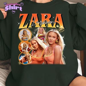 May include: Dark green sweatshirt featuring a graphic of Zara Larsson with the text "ZARA Larsson" in orange and white. The design includes multiple images of the singer, with a vintage aesthetic.