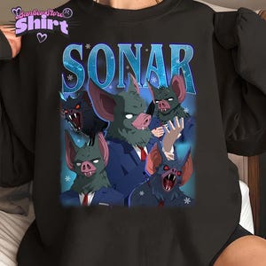 Puede incluir: Sudadera negra con un gráfico de criaturas parecidas a murciélagos con trajes, la palabra "SONAR" en azul y copos de nieve. El diseño tiene una estética vintage. La sudadera tiene cuello redondo.