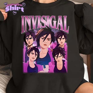May include: Black sweatshirt with a graphic of a woman in a pink jacket, and the word "INVISIGAL" in a pink and white font. The sweatshirt has a crew neck and long sleeves. The graphic is a vintage style design.