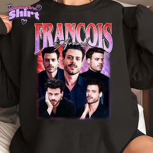 May include: Black sweatshirt with a graphic print of Francois Arnaud's image and name in a retro style. The design features multiple images of the actor against a purple and red backdrop. The sweatshirt has a crew neck and long sleeves.