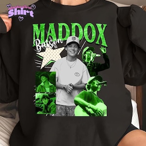 Puede incluir: Sudadera negra de cuello redondo con un diseño de collage que presenta a Maddox Batson. El diseño incluye el nombre "MADDOX" en verde, el nombre "Batson" en escritura blanca y varias imágenes del artista. También se incluye un gráfico de estrella.