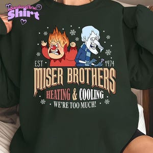May include: Dark green sweatshirt featuring a graphic of the Miser Brothers from the 1974 animated film. The design includes the text "Miser Brothers Heating & Cooling" and "We're Too Much!" with snowflake accents. The sweatshirt has a crew neck.