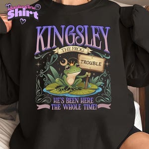 May include: Black t-shirt featuring a green frog wearing a crown and holding a sign that reads "Trouble". The text "Kingsley the Frog" is above the frog and the text "He's been here the whole time!" is below the frog.