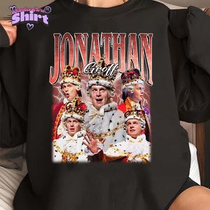 May include: Black sweatshirt featuring a graphic of Jonathan Groff in a royal costume with a crown. The name "Jonathan Groff" is printed in a vintage style with a red and white gradient. The shirt has the text "Panther Store Shirt" in the upper left corner.