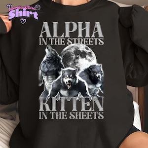 May include: Black sweatshirt featuring a graphic of three werewolves in front of a full moon. The text "ALPHA IN THE STREETS" and "KITTEN IN THE SHEETS" is printed above and below the image in a silver, textured font. The sweatshirt has a crew neck.