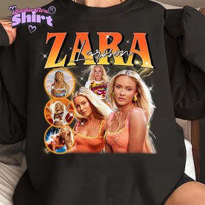 May include: Black sweatshirt featuring a graphic of Zara Larsson with the text "ZARA Larsson" in orange and white. The design includes multiple images of the singer with a retro aesthetic, and lightning bolt accents.