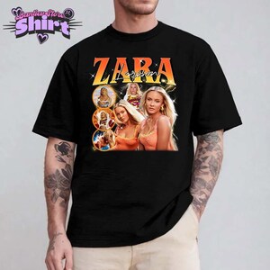 May include: Black t-shirt featuring a graphic of Zara Larsson with the name "ZARA" in orange and yellow lettering. The design includes multiple images of the singer. The shirt is a classic crew neck style.