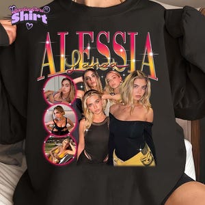 Pode incluir: Camisola preta com um gráfico retro do nome "Alessia Lanza" e um grupo de mulheres. O design incorpora três imagens circulares de uma mulher em várias poses. A camisola tem gola redonda.
