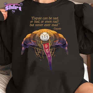 Puede incluir: Camiseta negra con un gráfico de una criatura mecánica estilizada y una pancarta con símbolos. La camiseta también tiene la cita: "Esquie can be sad, or bad, or even rad! but never ever mad" y el nombre "-Esquie".