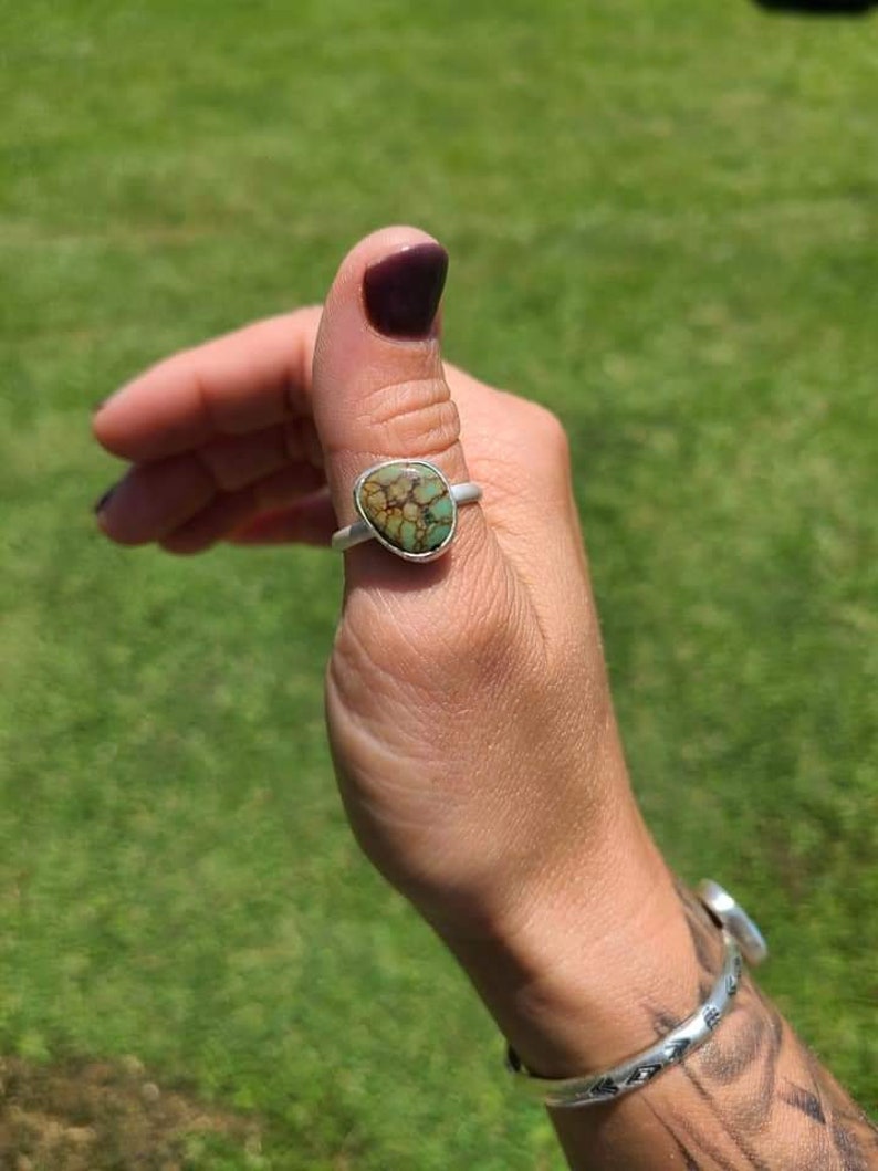 Op de afbeelding: Een zilveren ring met een groene en bruine steen in het midden. De ring wordt op de duim gedragen.