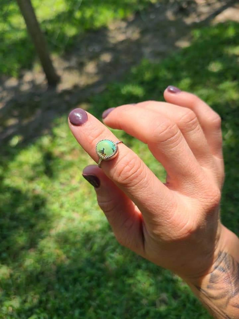 May include: A silver ring with a green gemstone set in a bezel setting. The ring is on a finger with dark purple nail polish.