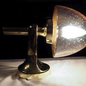 Op de afbeelding: Een gouden wandlamp met een getextureerde glazen kap. Het licht is aan en de gloeilamp is zichtbaar.