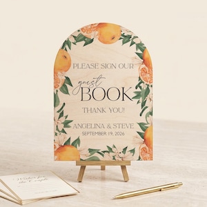 May include: Wooden guest book sign with an arch top, featuring orange and floral illustrations. The sign reads "PLEASE SIGN OUR guest BOOK THANK YOU!" with the names Angelina & Steve and the date September 19, 2026.