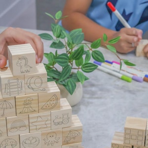May include: Wooden building blocks with various black line drawings of objects and animals, including a zebra, rainbow, and pizza. A hand is placing a block on top of the stack. Colored markers and a small plant are also visible.