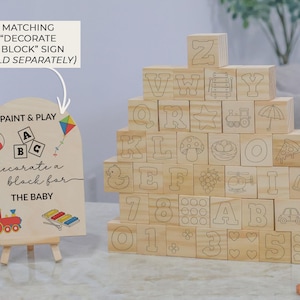 May include: Wooden alphabet blocks with etched letters, numbers, and illustrations, stacked in a pyramid. A wooden sign with the text "Paint & Play" and "decorate a block for THE BABY" is in front of the blocks. The sign has a colorful kite and other illustrations.