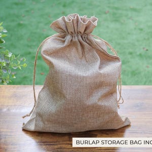 May include: A beige burlap drawstring bag with a gathered top. The bag is sitting on a wooden surface with a green background. The text "BURLAP STORAGE BAG INCLUDED" is at the bottom.