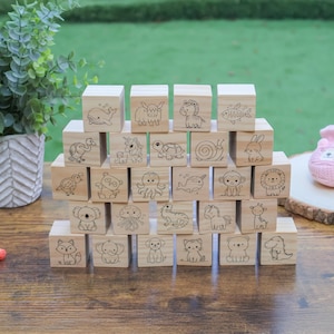 May include: A stack of wooden blocks with engraved animal illustrations, including a whale, owl, and zebra. The blocks are arranged on a wooden surface, with colorful markers and pink baby booties nearby. A potted plant is in the background.